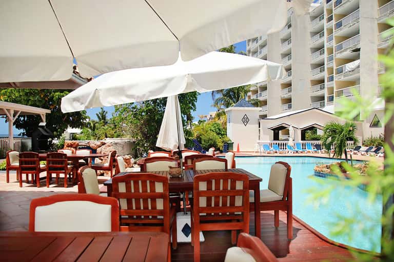 Poolside dining area with red wooden chairs, white umbrellas, and multi-story resort building in background