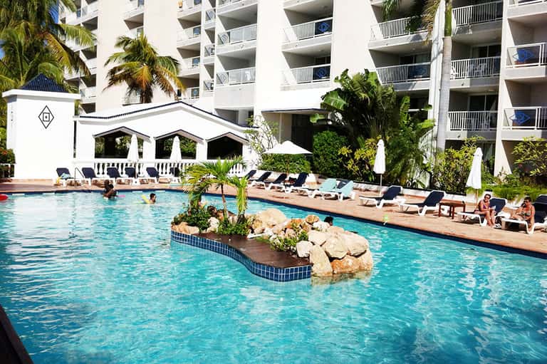 Resort pool with island centerpiece, palm trees, and multi-story hotel building backdrop