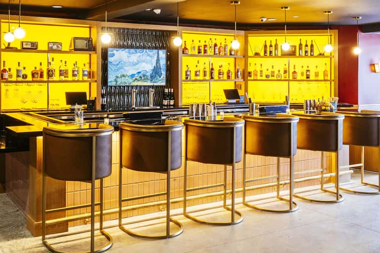 Modern bar with bright yellow shelving displaying liquor bottles, brown bar stools, and pendant lighting
