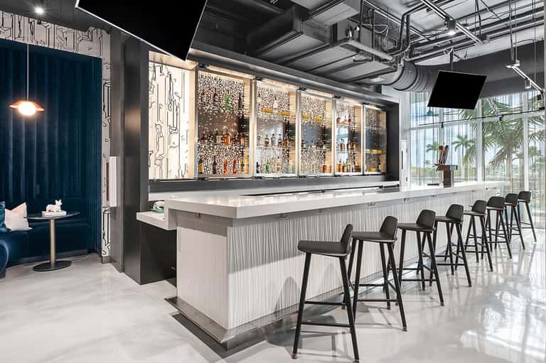 Modern hotel bar with illuminated glass shelving, white bar counter, black bar stools, and industrial ceiling