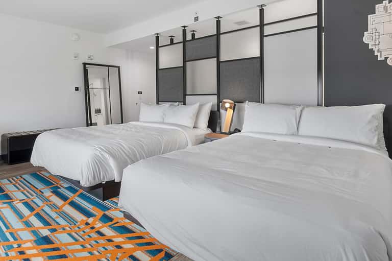 Contemporary hotel room with two queen beds, dark headboard, geometric patterned floor rug, and minimalist design