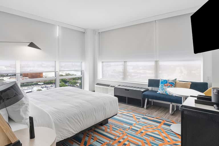 Modern hotel room with white bedding, blue sofa, expansive city views, and geometric area rug