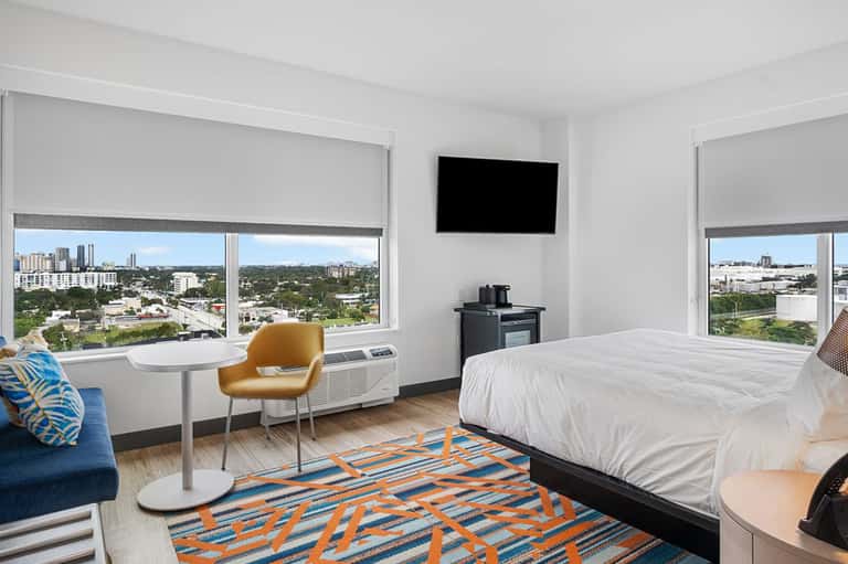 Modern hotel room with city skyline views, queen bed, desk, and contemporary furnishings