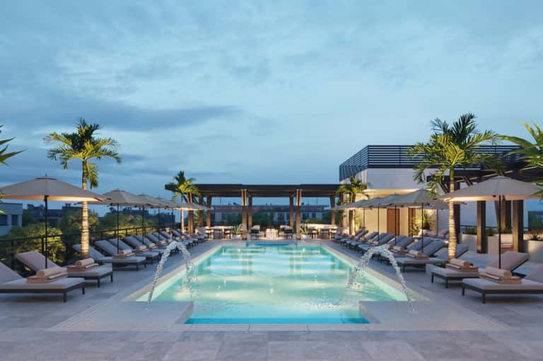 Luxury rooftop pool at dusk with loungers, palm trees, modern architecture, and city skyline views