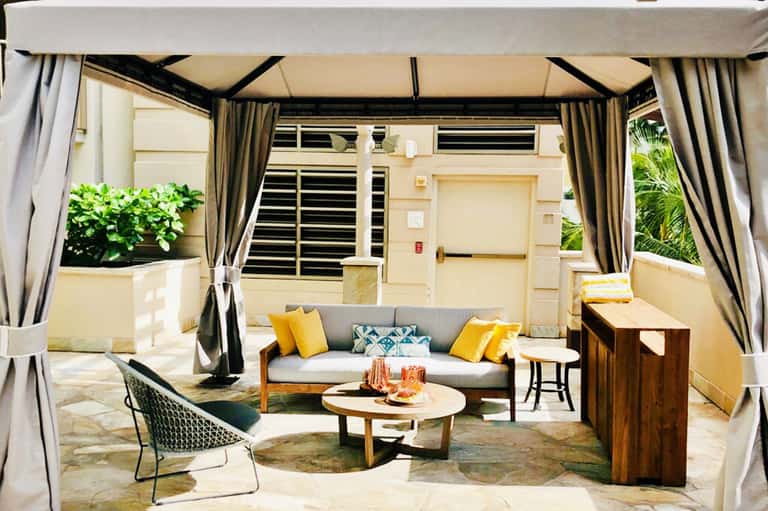 Covered patio lounge with gray sofa, yellow pillows, wooden tables, and draped curtains overlooking a building entrance