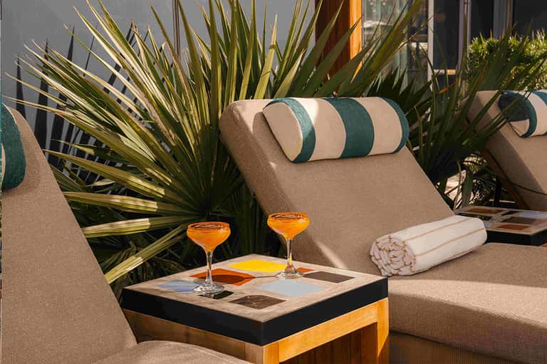 Outdoor lounge area with beige chairs, striped pillows, tropical plants, and cocktails on side table