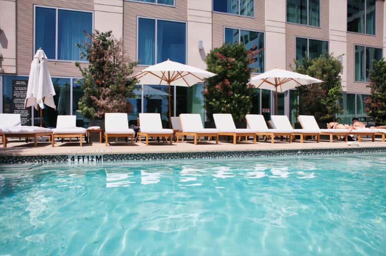 Resort pool with turquoise water, wooden loungers with white cushions, white umbrellas, and modern brick building backdrop