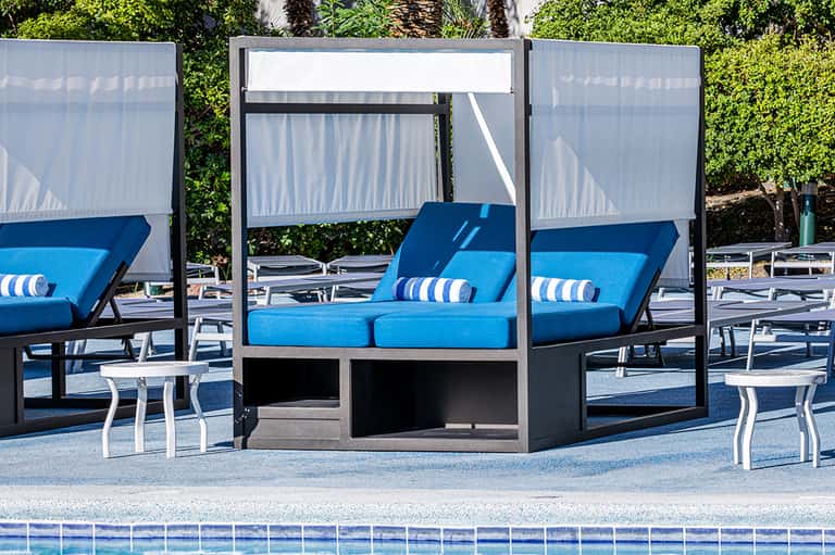 Poolside lounge area with blue cushioned daybed, white retractable shade, and green landscaping