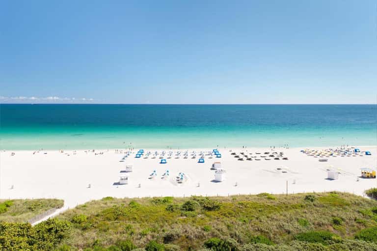 Aerial view of a crowded beach with turquoise water, white sand, and blue beach umbrellas
