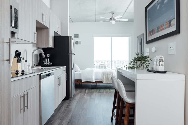 Compact studio hotel room with kitchenette, white bedding, desk area, and floor-to-ceiling windows.