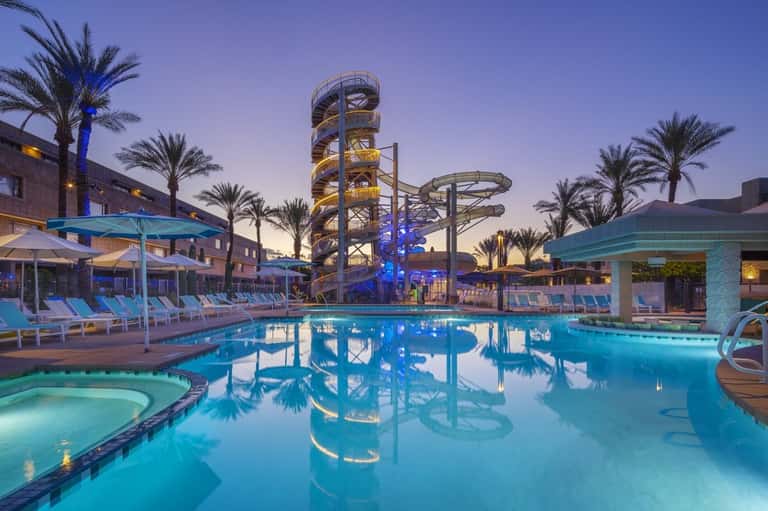 Illuminated resort pool complex at dusk with water slides, palm trees, and lounge chairs
