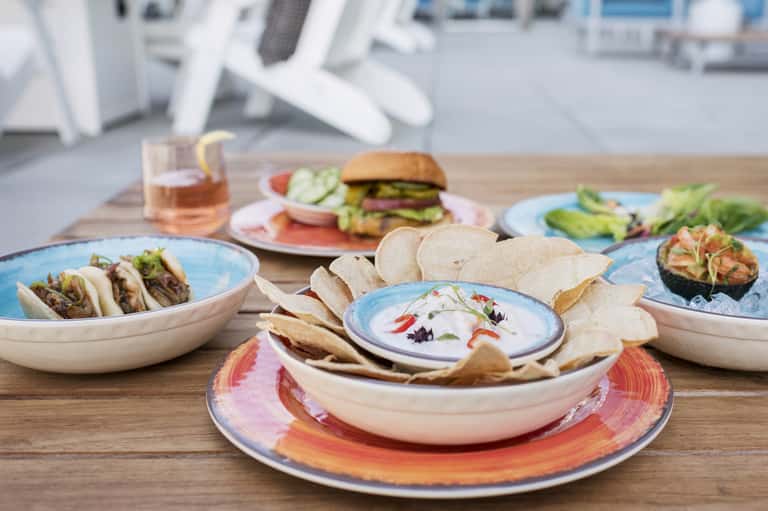 Beachside dining table with colorful plates of tacos, appetizers, and fresh drinks on wooden deck