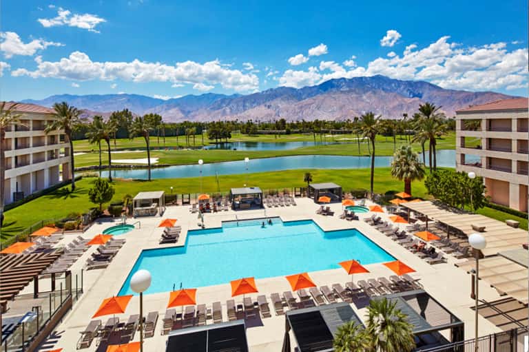 Luxury resort pool complex with mountain views, palm trees, and multiple lounging areas with orange umbrellas