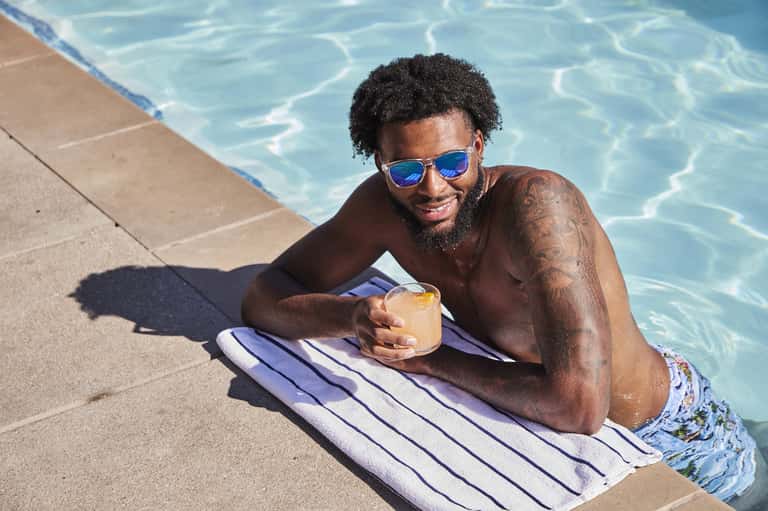 Man in blue sunglasses relaxing poolside with drink on striped towel