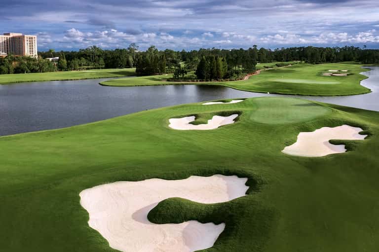 Championship golf course with sand bunkers, water hazards, lush fairways, and tree line under blue sky