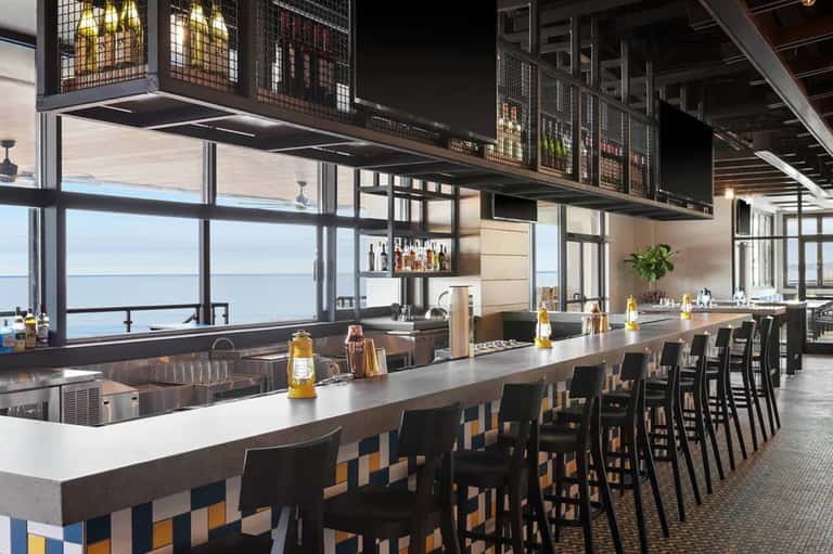 Contemporary bar with ocean views, white countertop, black stools, and bottles displayed on shelves overhead