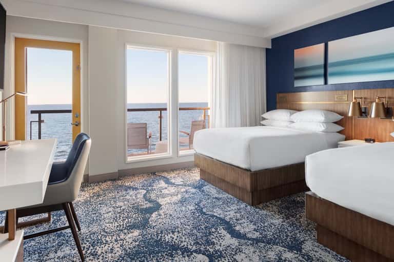 Modern hotel bedroom with ocean views, queen beds, desk, and glass balcony doors