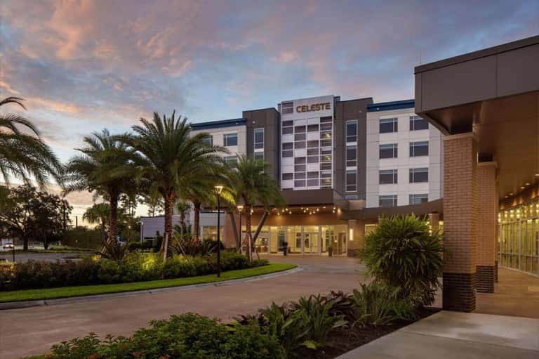 Hotel exterior at dusk with palm trees, brick facade, and illuminated entrance under purple sky