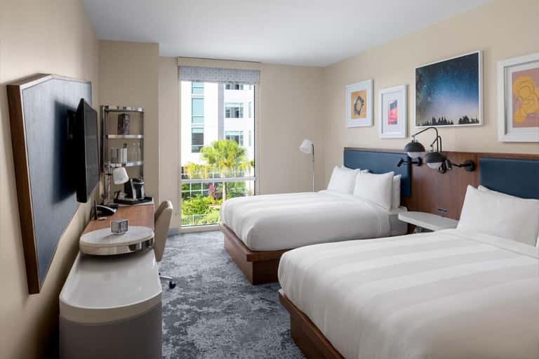 Bright hotel room with two queen beds, blue headboards, balcony view, flat-screen TV, and contemporary furnishings