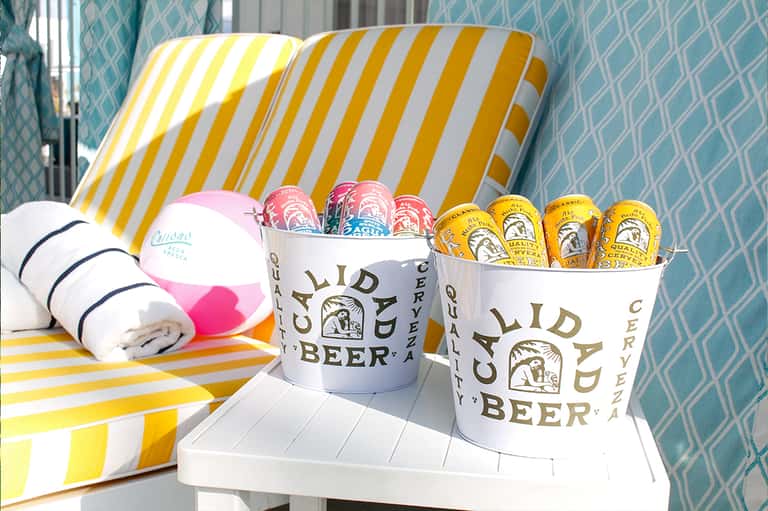 Beach lounge chair with yellow and white stripes, beer branded buckets, towels, and pink beach ball on sand