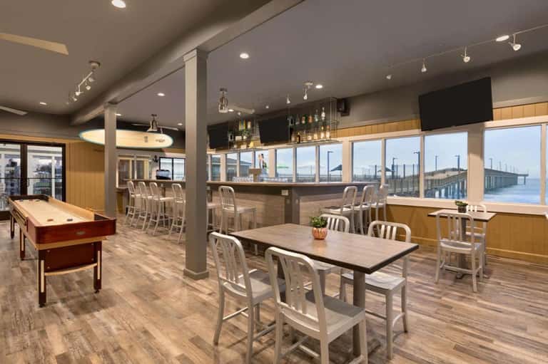 Modern indoor lounge with pool table, bar seating, floor-to-ceiling windows overlooking water and industrial beam ceiling