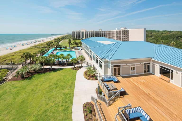 Beachfront resort with multi-story hotel building, curved pool with palm trees, wooden deck area, and beach access on sunny day