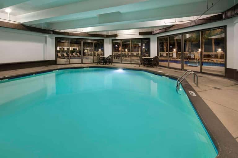 Indoor swimming pool with turquoise water, ceiling beams, lounge chairs, and glass doors to outdoor patio