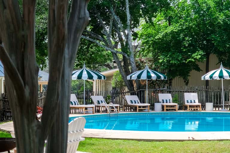 Resort pool with striped umbrellas and lounge chairs surrounded by mature trees and wooden deck