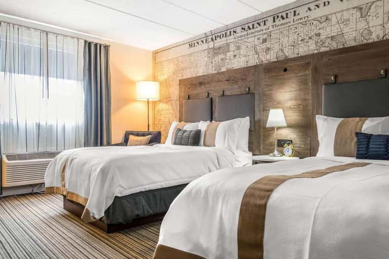 Modern hotel room with two queen beds, wood headboards, and vintage map wall décor in warm lighting