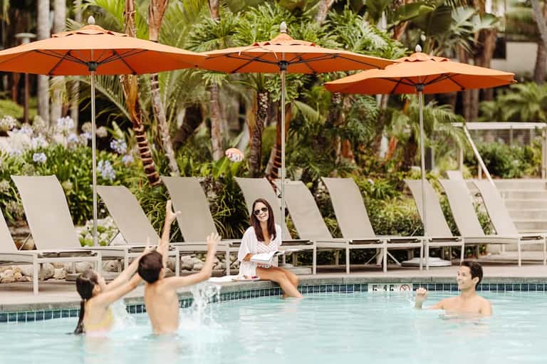 Resort pool with guests swimming, orange umbrellas, and lounge chairs with tropical landscaping