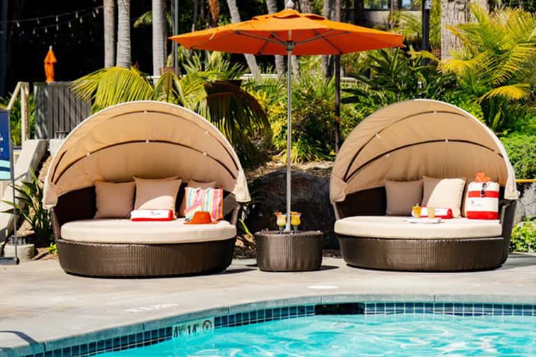 Two beige dome-shaped poolside cabanas with an orange umbrella and cushioned seating beside a swimming pool