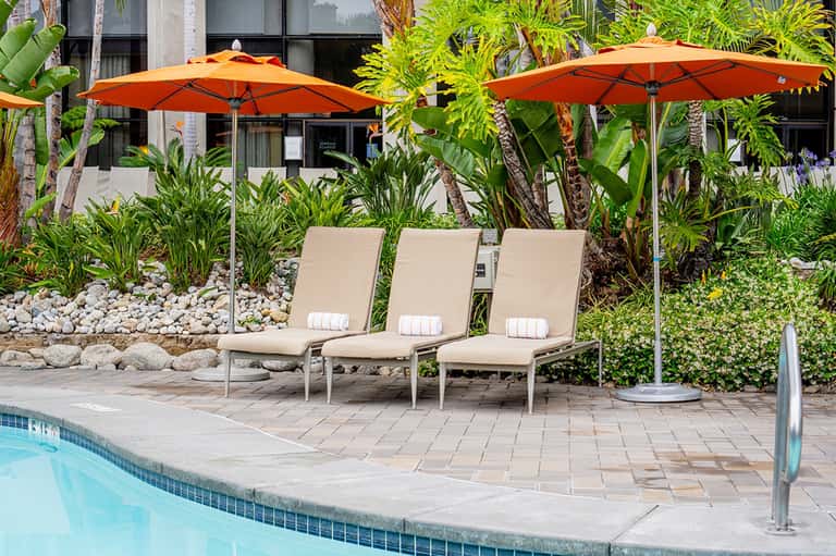 Poolside lounge area with three tan chairs under orange umbrellas surrounded by tropical plants
