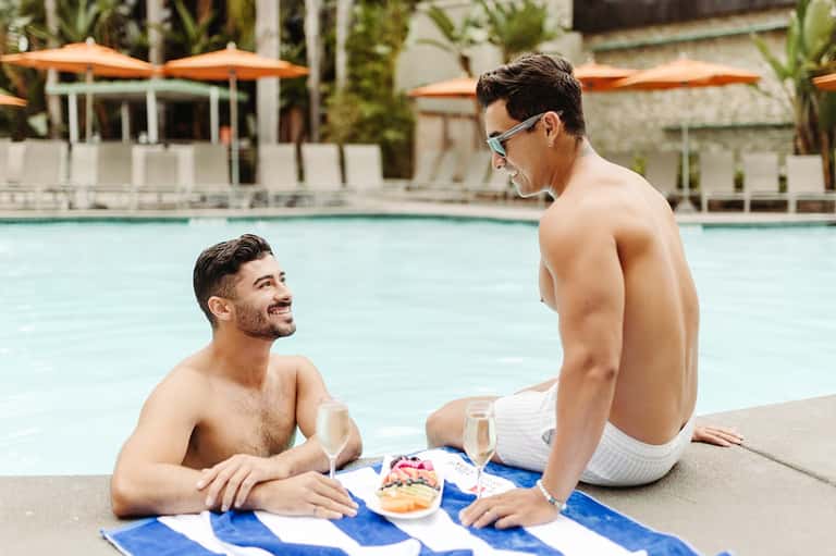 Two men sitting poolside enjoying beverages and snacks by a resort pool with umbrellas