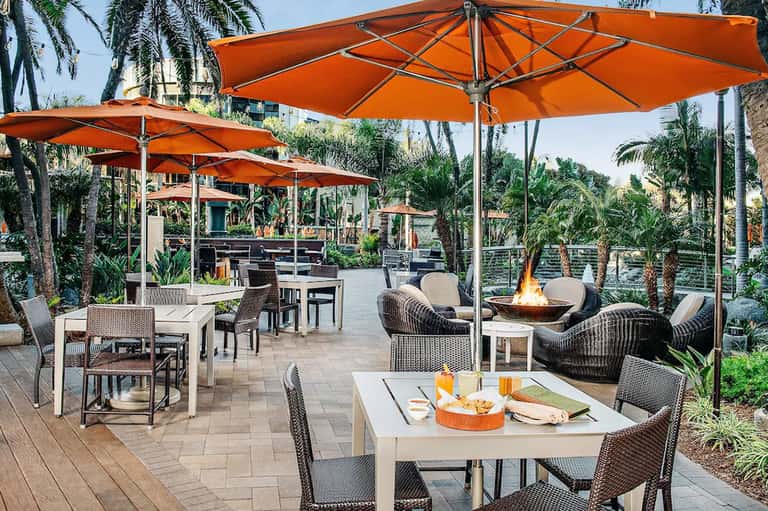 Outdoor patio with orange umbrellas, wicker seating, fire pit, and palm trees at dusk