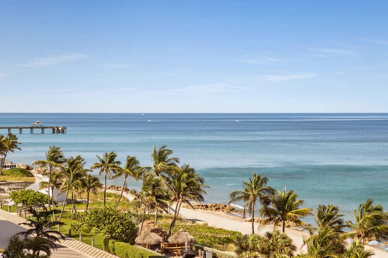Tropical beachfront resort with palm trees, white sand beach, and ocean pier visible in the distance