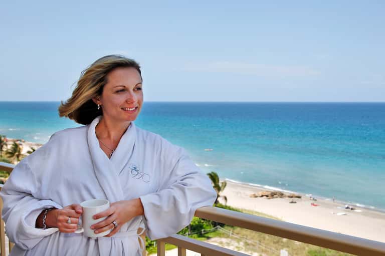 Woman in white spa robe holding coffee cup on oceanfront balcony overlooking beach