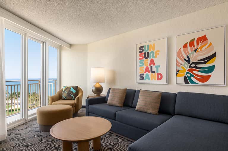 Coastal hotel suite with ocean view, modern sofa, colorful wall art, and balcony access