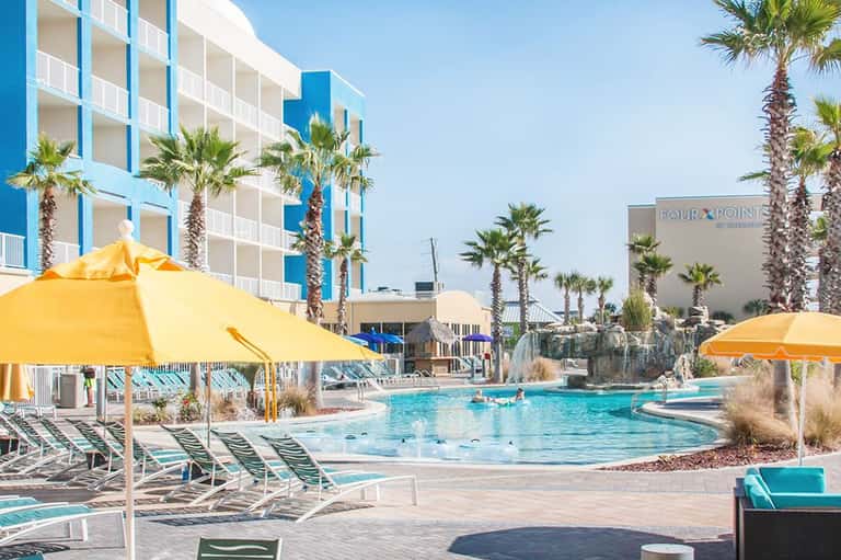 Resort pool with turquoise water, yellow umbrellas, lounge chairs, and blue high-rise hotel building with palm trees