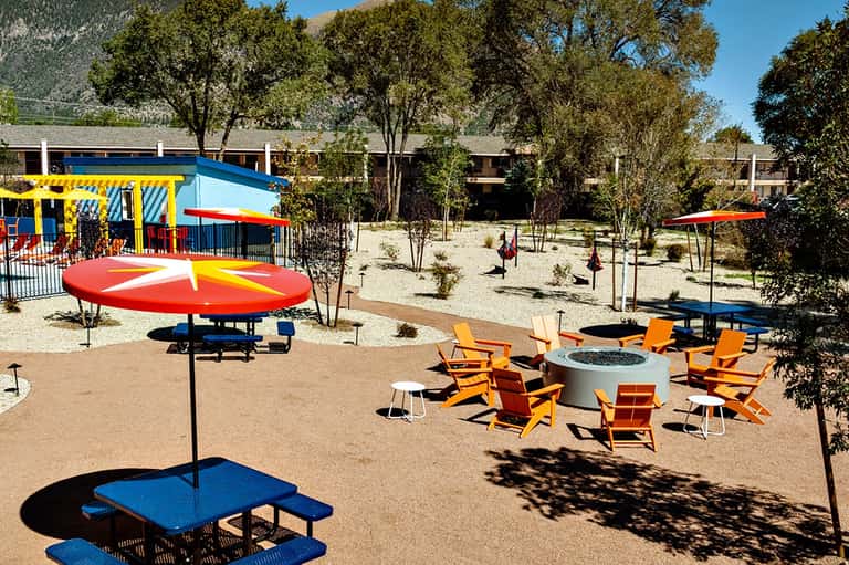 Colorful outdoor recreation area with playground equipment, fire pit seating, and shade umbrellas