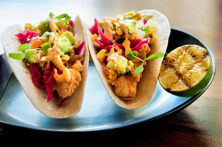 Two gourmet fish tacos with colorful toppings and grilled lime on white plate