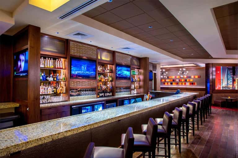 Modern bar with wooden cabinetry, multiple TV screens, granite countertop, and purple-upholstered bar stools