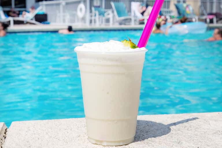 Frozen cocktail with whipped cream and pink straw by resort pool with swimmers