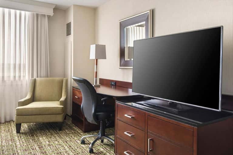 Modern hotel room with work desk, large TV, green armchair, wood furniture, and neutral décor