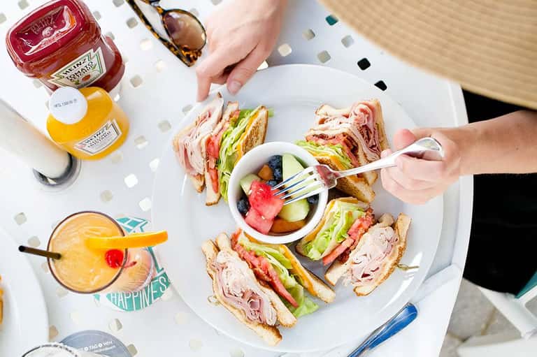 Person eating sandwiches with fresh fruit bowl and cocktails on white plate at outdoor dining table