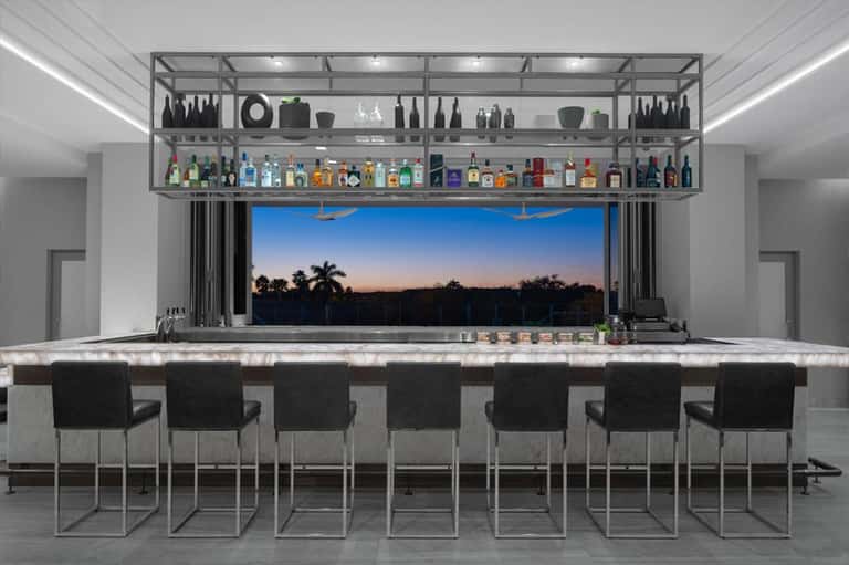 Upscale home bar with metal shelving, marble countertop, black bar chairs, and panoramic window view at dusk
