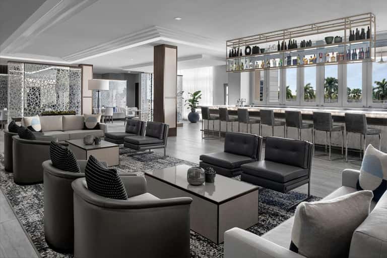 Luxury lounge with modern furniture, bar counter, and floor-to-ceiling windows overlooking palm trees