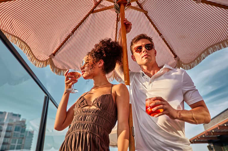 Couple enjoying cocktails under a striped umbrella on a rooftop terrace with city skyline