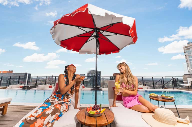 Two women enjoying drinks under red and white umbrella by rooftop pool with city skyline