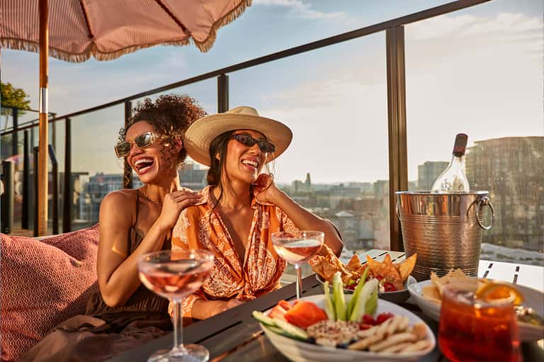 Two women enjoying cocktails on a rooftop terrace with city skyline views, appetizers, and wine bucket