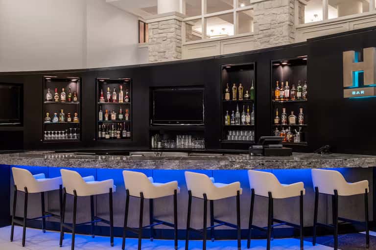 Contemporary bar with black backing, illuminated shelving displaying spirits, and cream upholstered bar stools